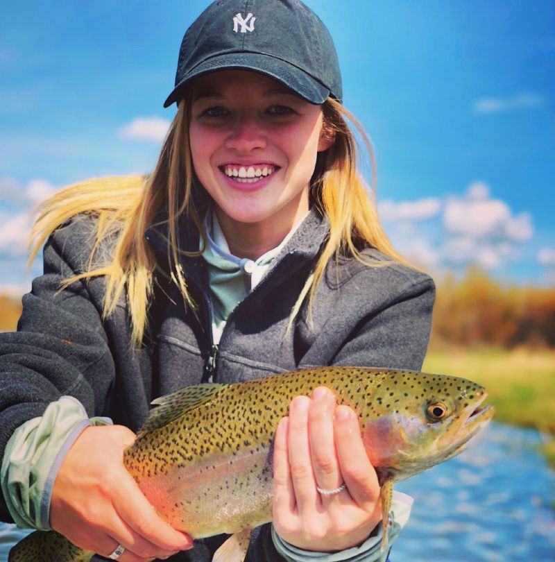 Woman smiling and holding a speckled fish by a blue lake on a sunny day