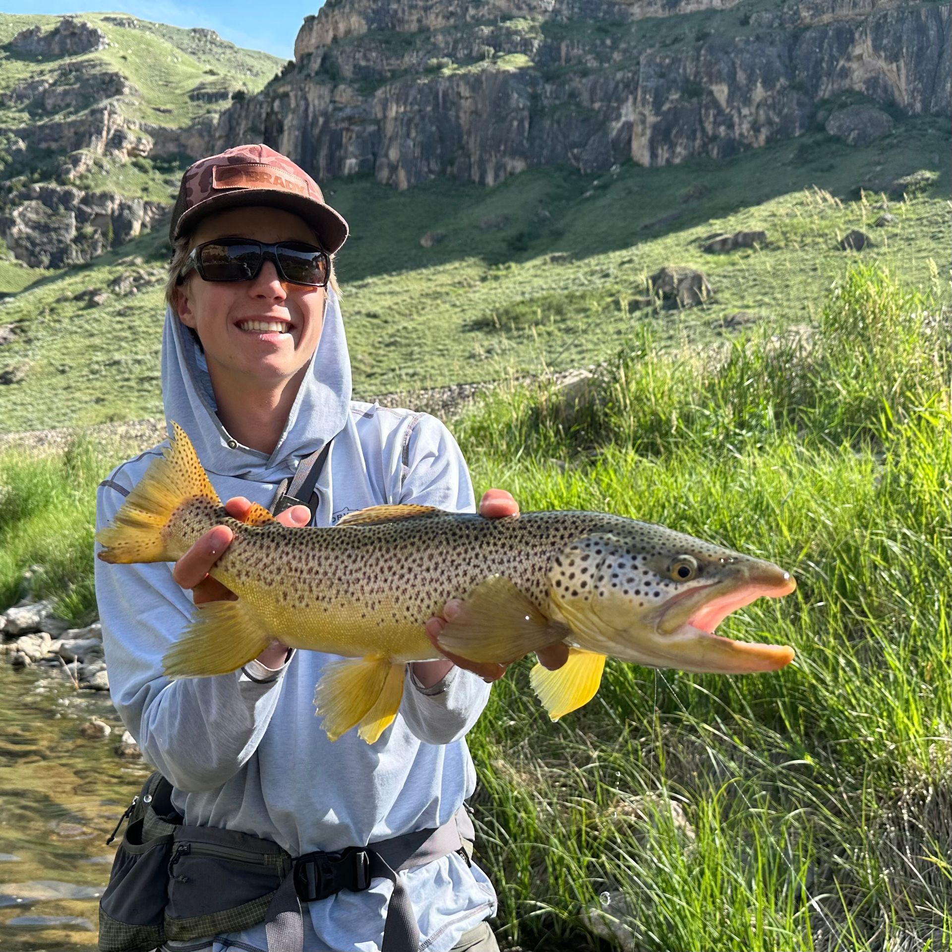 Smiling angler holding a large trout by a grassy mountain stream under sunny skies