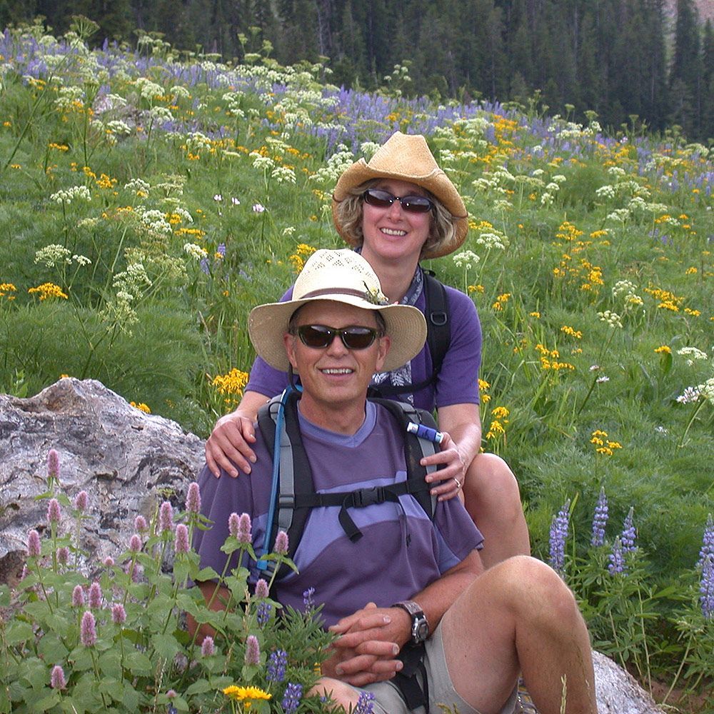 Two smiling hikers in sun hats sitting among wildflowers on a mountain meadow