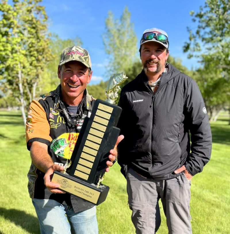 Two smiling men outdoors holding a tall trophy on a sunny lawn