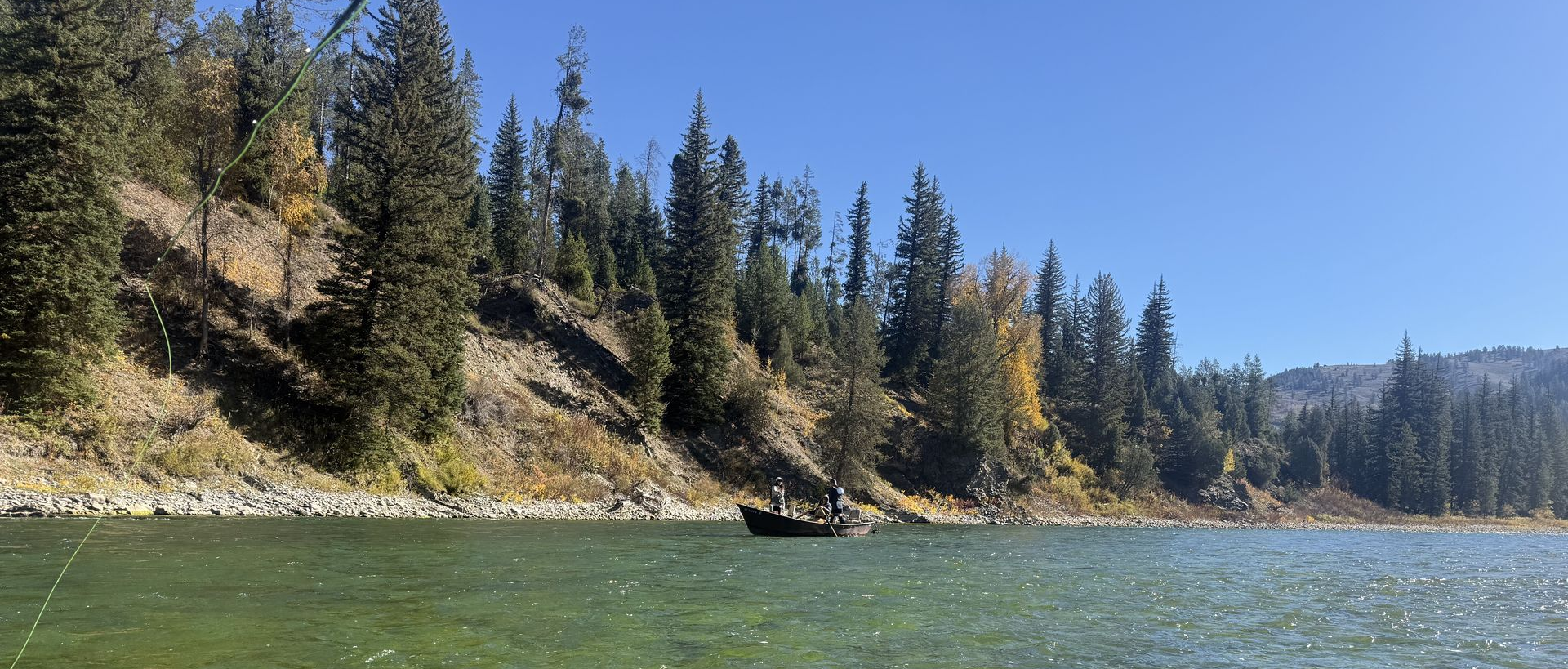 Person in black and green jacket by a river, holding a camera toward a rocky forested canyon.