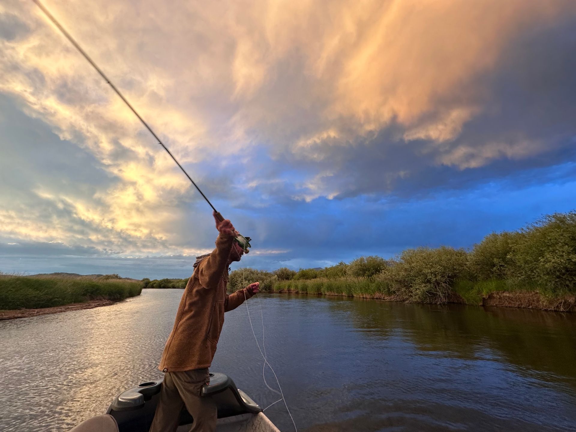 Person fly-fishing in a shallow river with a forested bank in the background