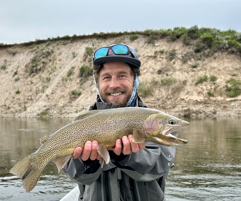 Man holding a large trout by a river, smiling, with rocky banks in the background