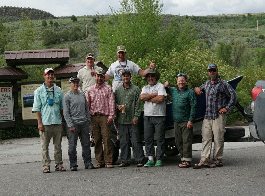 Group of hikers posing outdoors beside a trailhead sign and parked vehicles, with green hills behind them