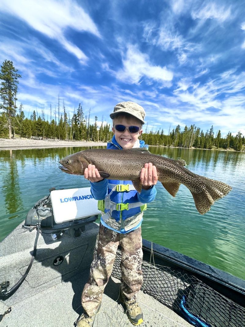 Child holding a large fish on a boat on a calm lake with pine trees and a blue sky