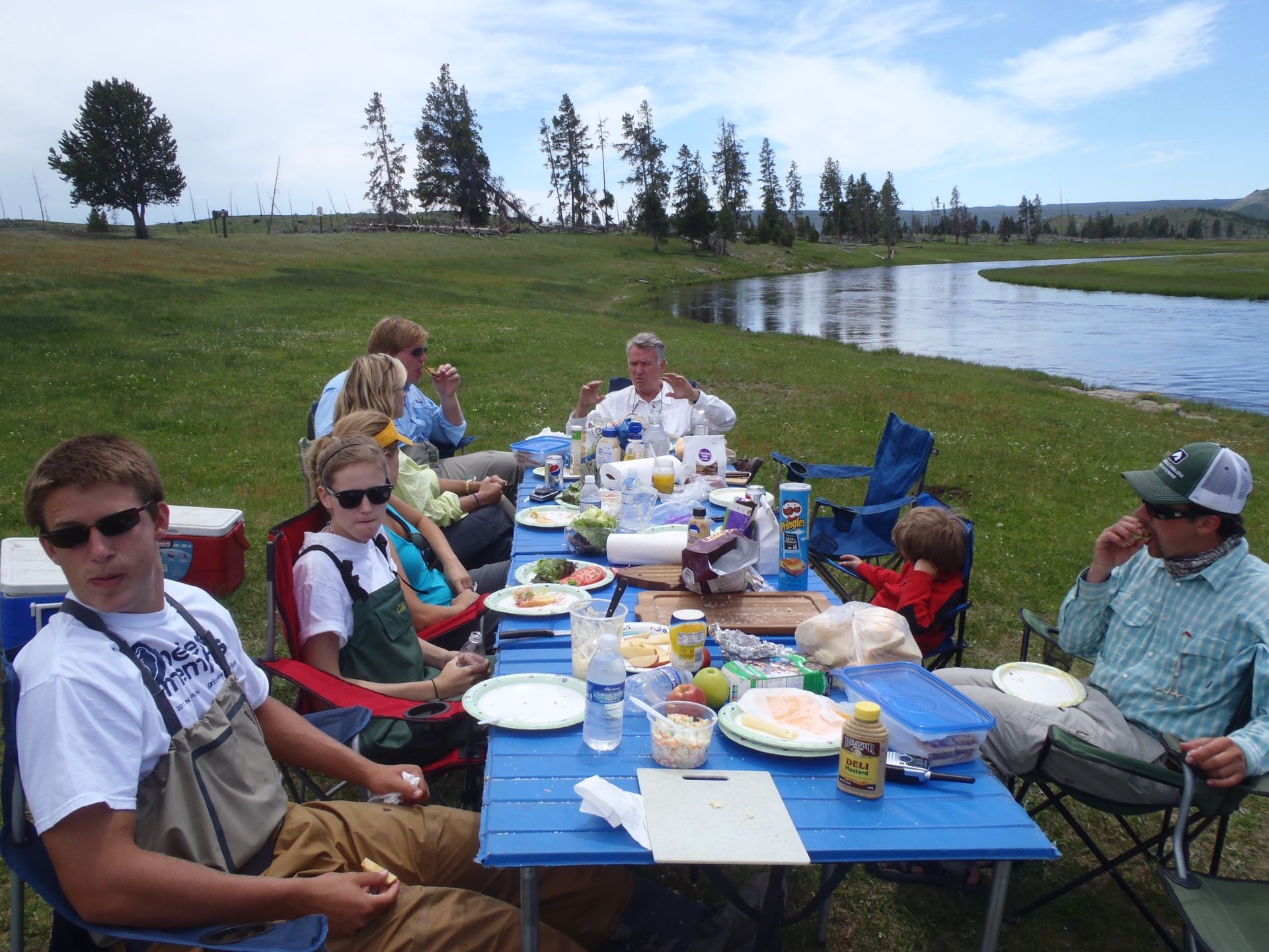 People seated at a blue picnic table by a lake, sharing food outdoors on a sunny day.