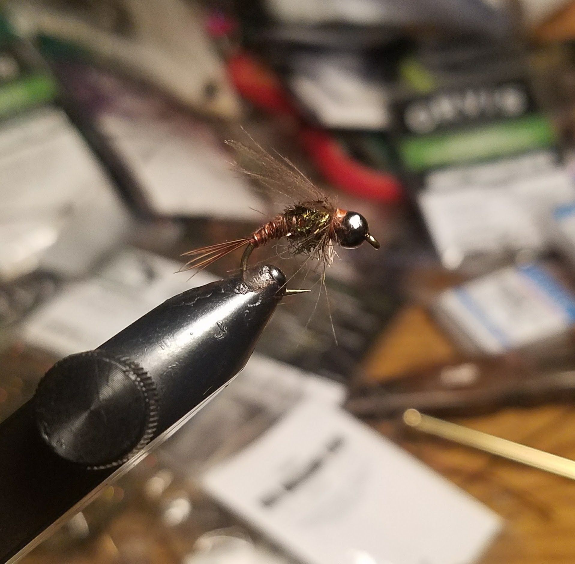Small brown fishing fly tied in a vise, with a blurred workbench background.