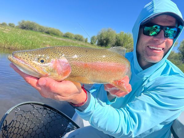 Smiling angler holding a large trout beside a net on a sunny riverbank