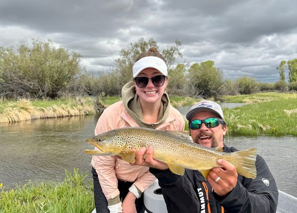 Two anglers hold a large golden fish by a river under cloudy skies.