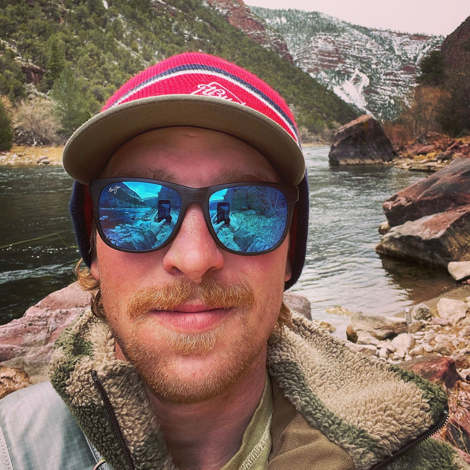 Selfie of a person in sunglasses and a striped hat by a rocky river with mountains behind