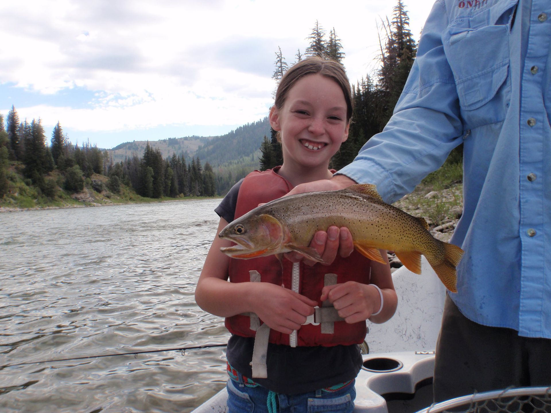 Smiling child holding a fish on a riverbank beside an adult, with mountains and trees in the background
