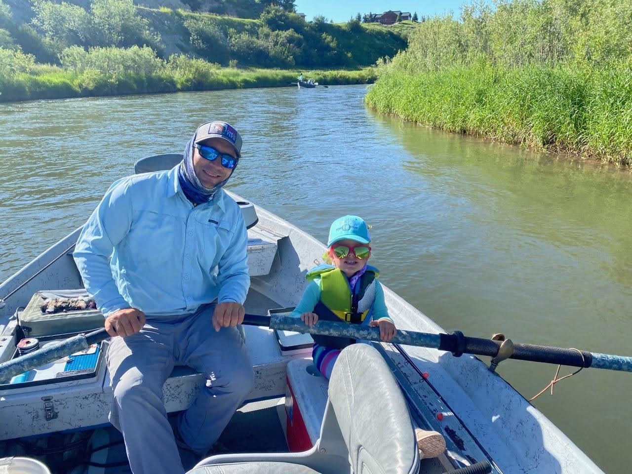 Two people fishing in a small boat on a calm river, with green banks and blue sky.