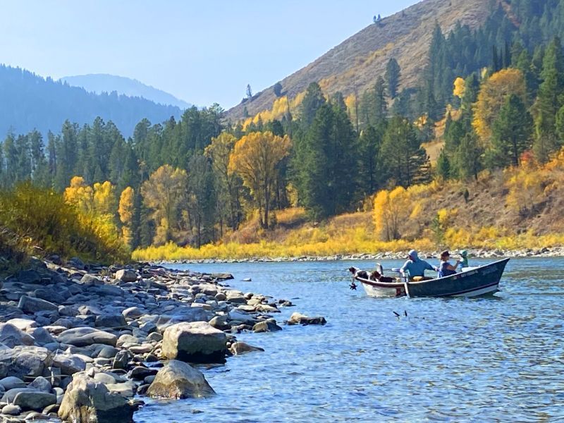 Two people in a small boat on a blue river beside rocky shore and autumn forested hills.