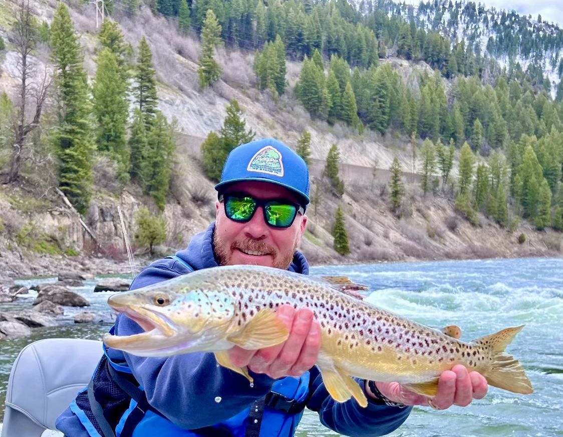 Angler holding a speckled trout by a mountain river with forested hills in the background