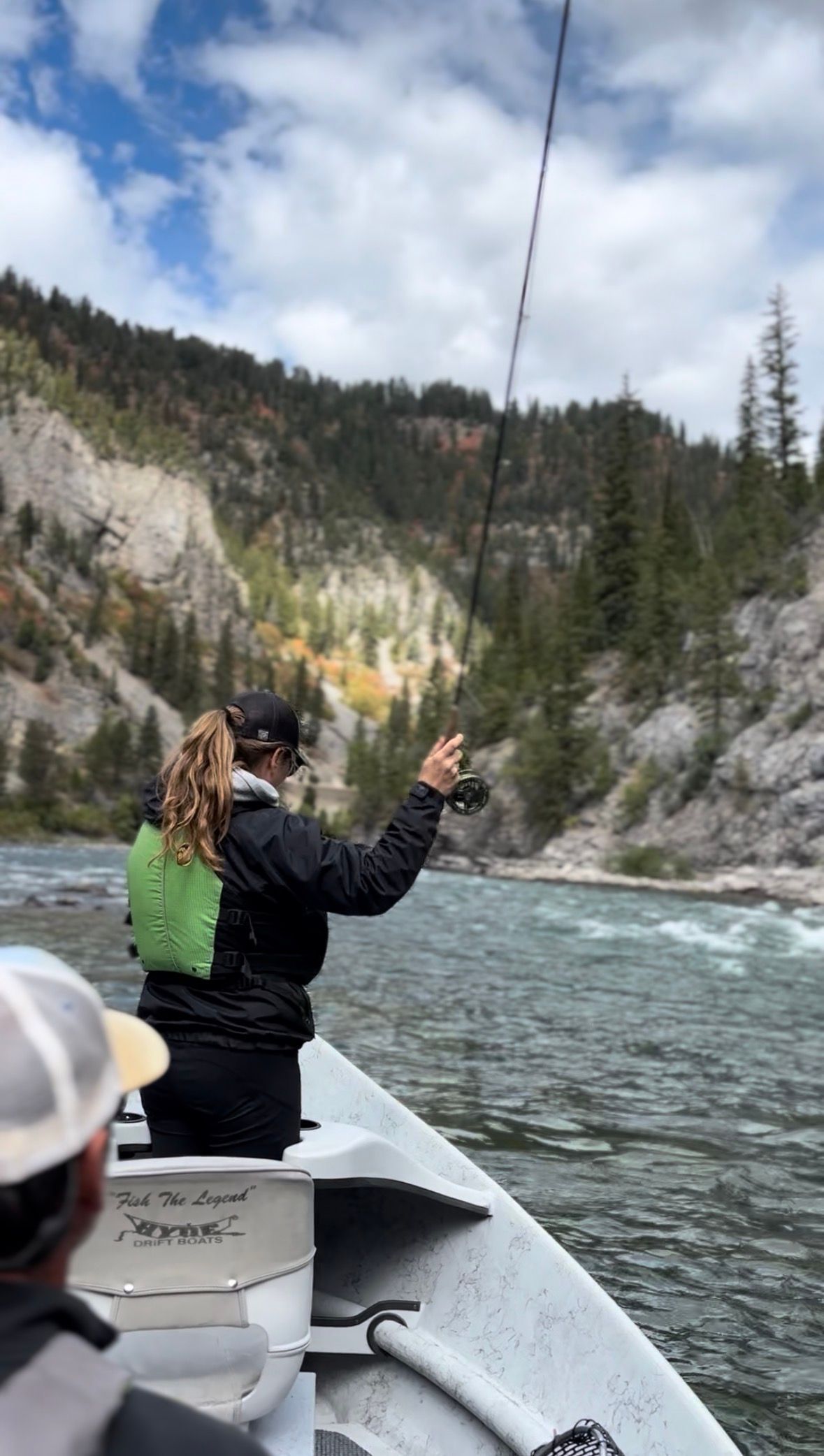 Person in a canoe pointing at rocky riverbanks and pine trees under a cloudy sky.