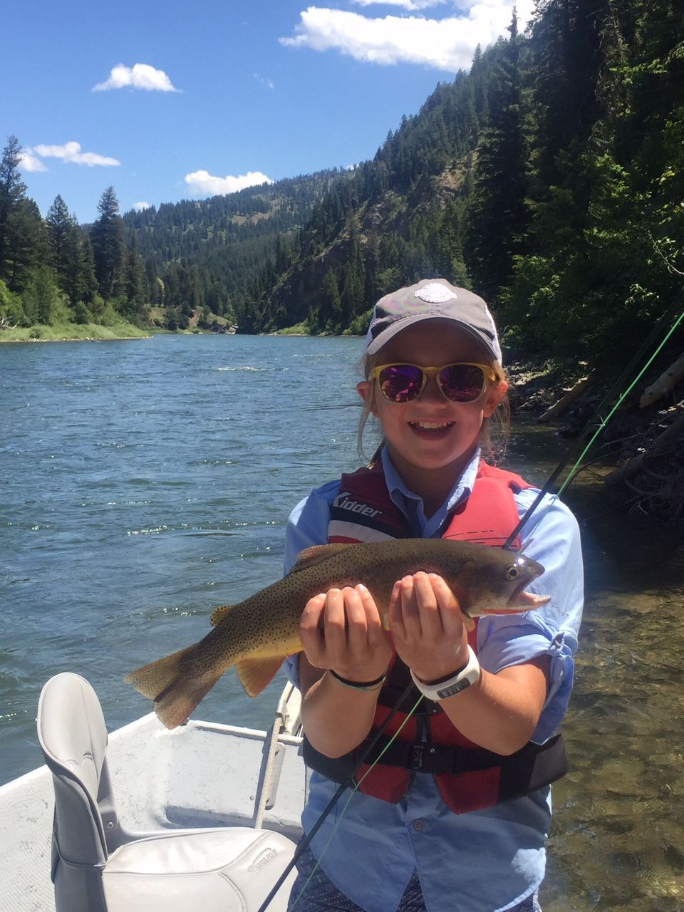Person in a boat on a river holding a fish, with forested mountains and blue sky in the background