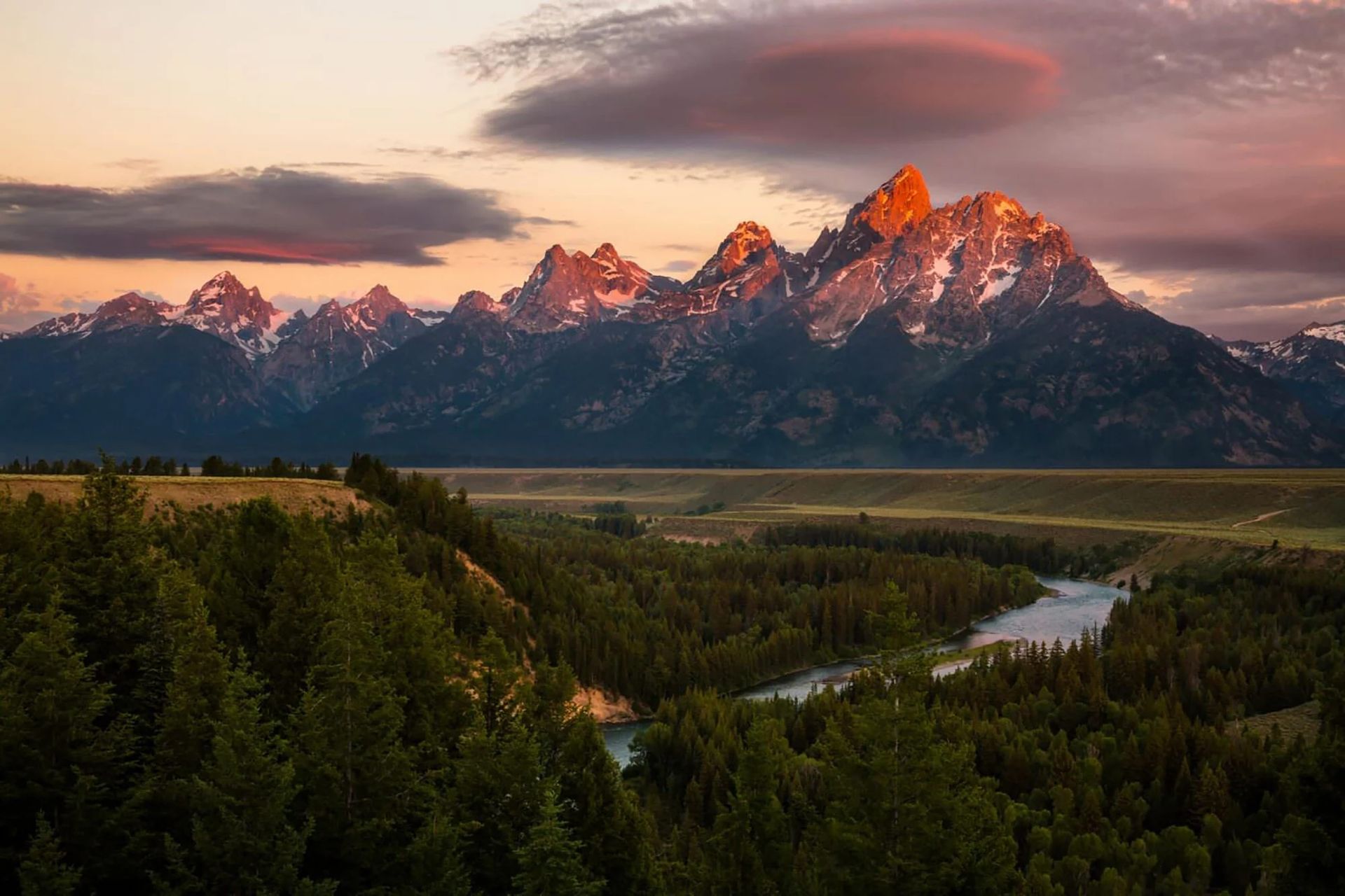 Snow-capped mountains at sunset above a winding river and evergreen forest
