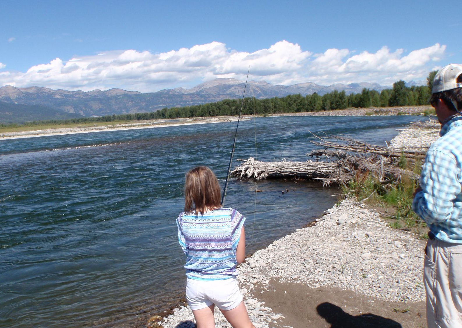 Two people standing by a river shore, looking at the water with mountains in the background.