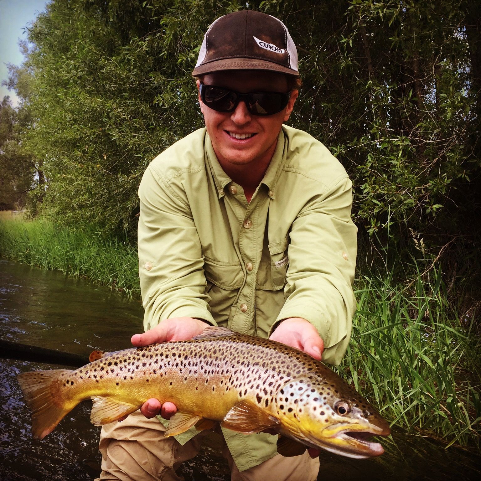 Angler holding a brown-speckled trout beside a river, wearing sunglasses and a cap