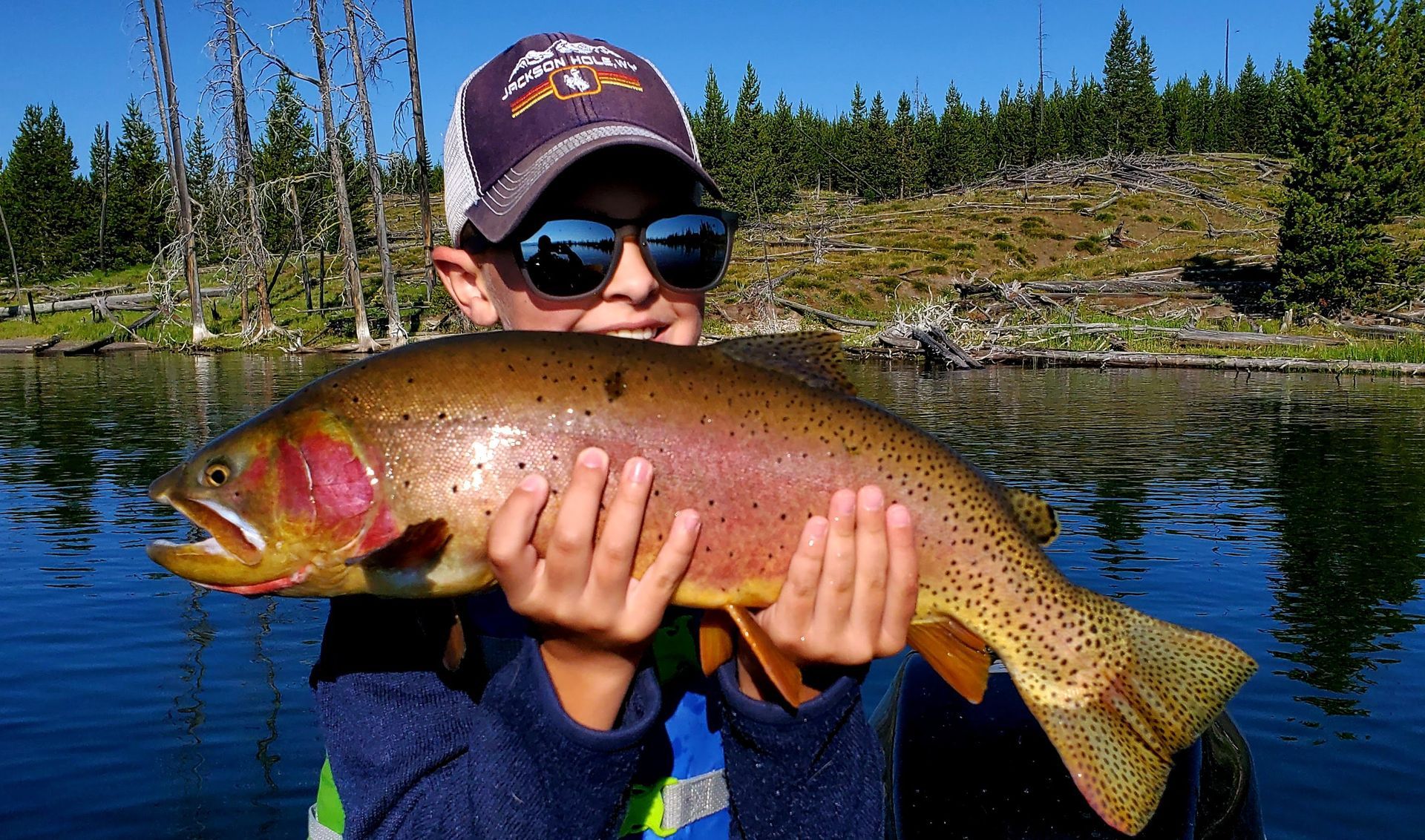 Person holding a large colorful fish by a calm lake with pine trees in the background