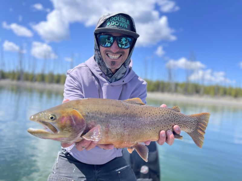 Angler smiling and holding a large rainbow trout beside a calm lake under a blue sky