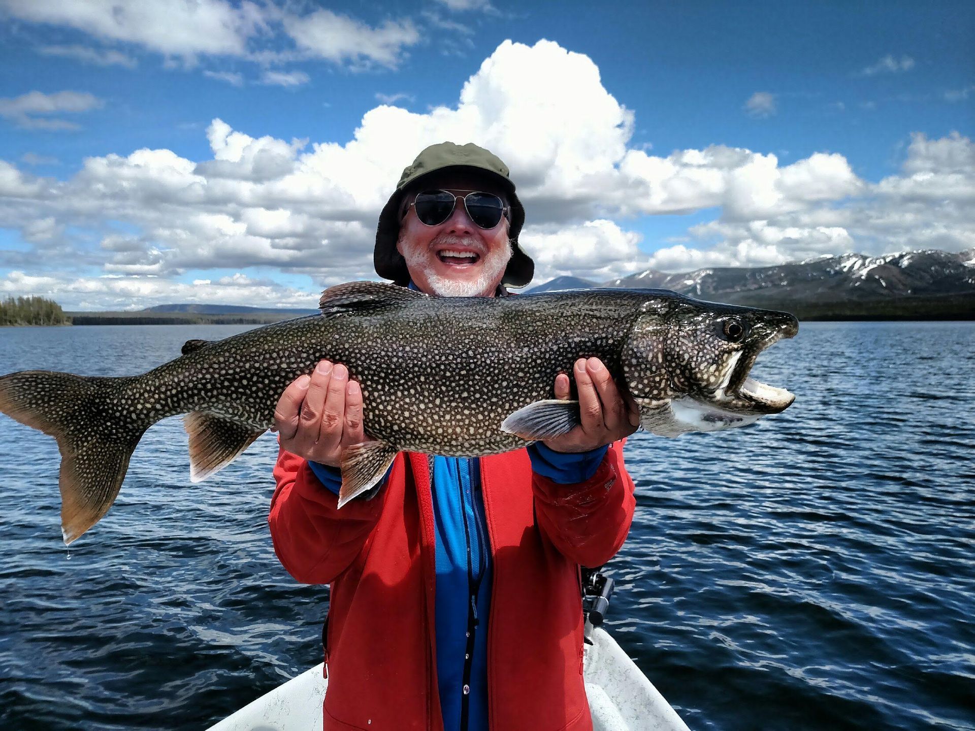 Angler in red jacket holding a large fish on a boat with mountains and water in the background