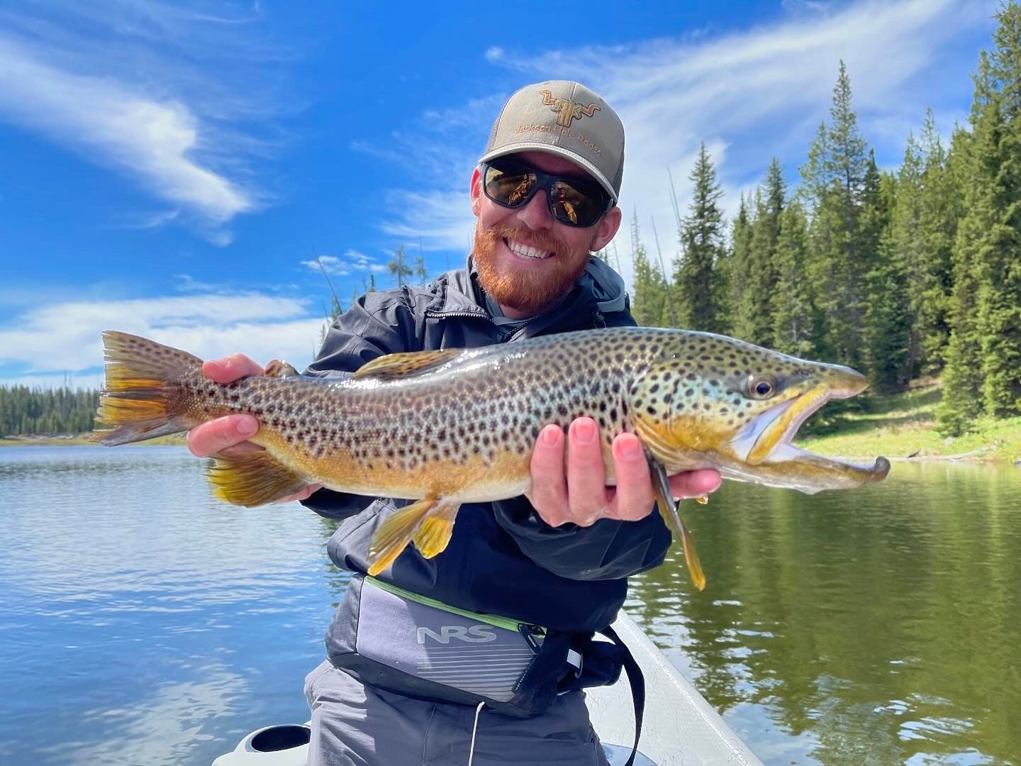 Angler holding a large spotted trout beside a calm lake with pine trees and blue sky.
