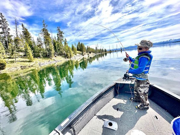 Person fishing from a boat on a calm lake beside a pine-lined shore under a cloudy blue sky