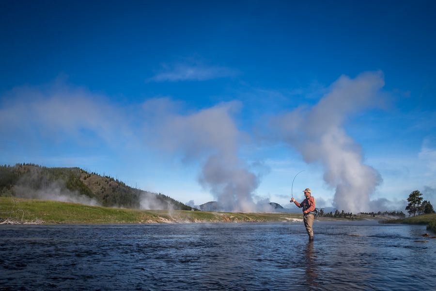 Person fly-fishing in a river with steam rising from geysers under a blue sky