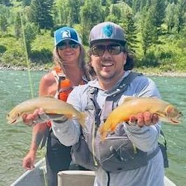 Two anglers holding two fish on a river, with a forested mountain backdrop