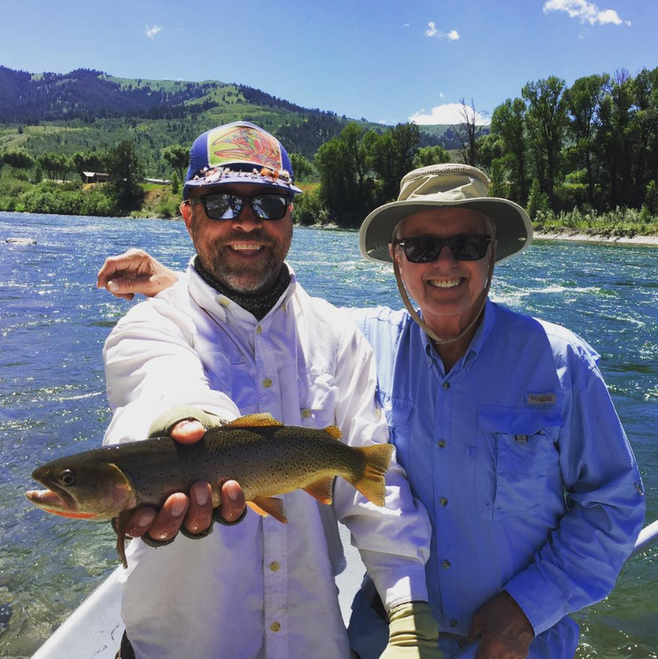 Two anglers by a river, smiling and holding a trout against a mountain backdrop