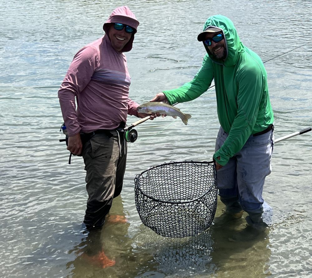 Two anglers in sun hats and hoodies holding a fish and a net in shallow water
