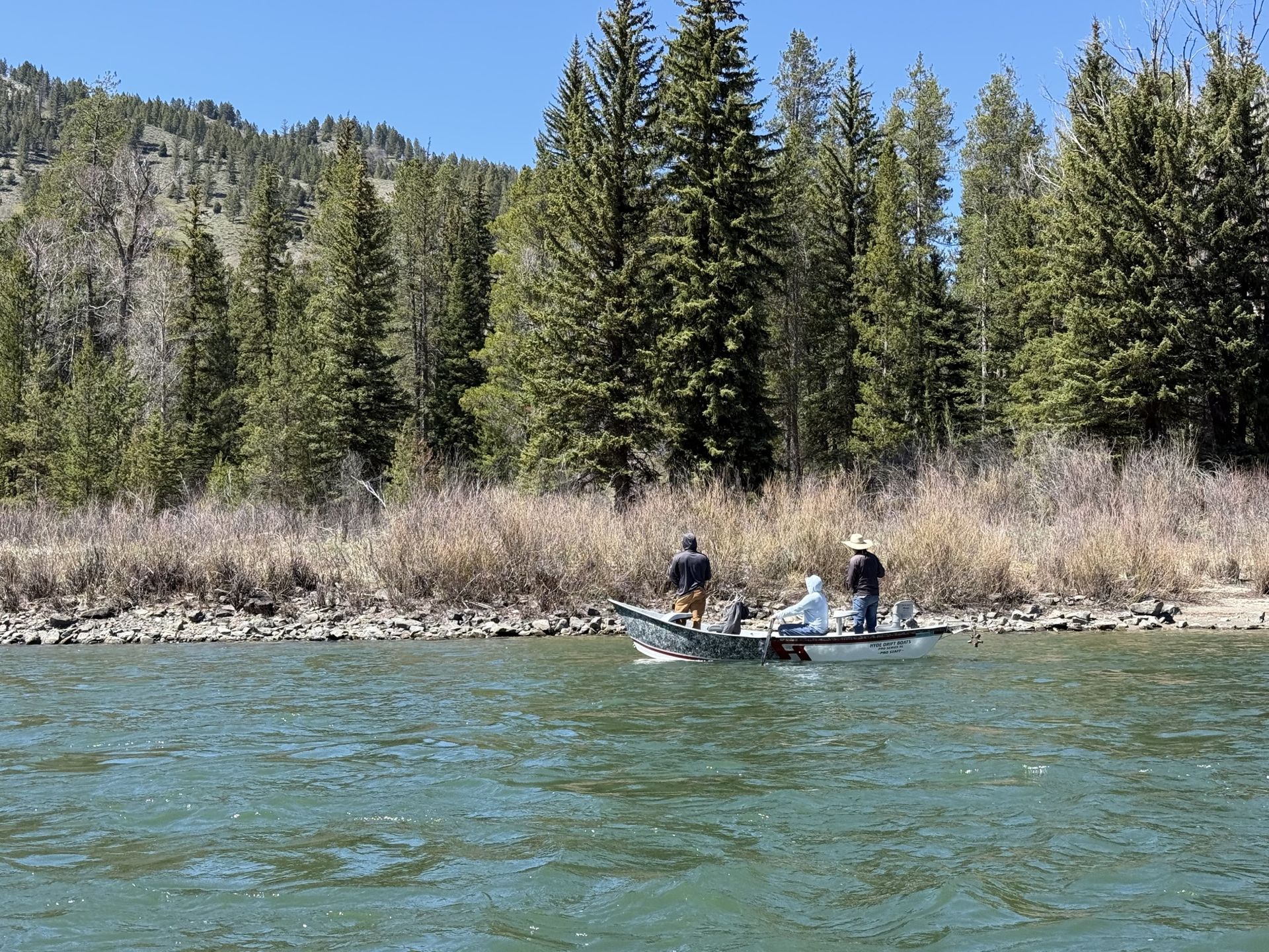 Two people in a boat on a blue river with pine trees and rocky banks in the background