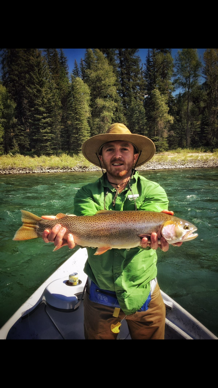 Man holding a large fish on a boat, wearing a straw hat and green shirt on a river with forest behind
