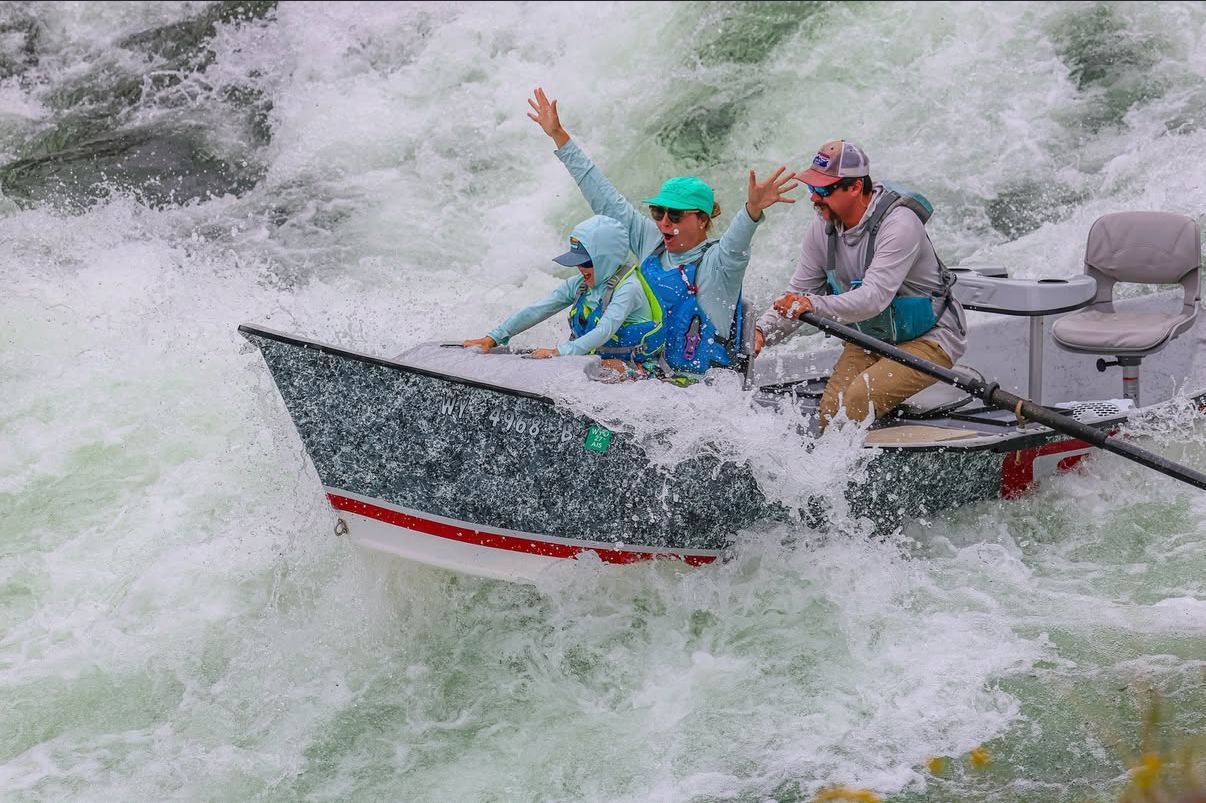Two people in a small boat bracing through churning whitewater rapids, spraying water all around.