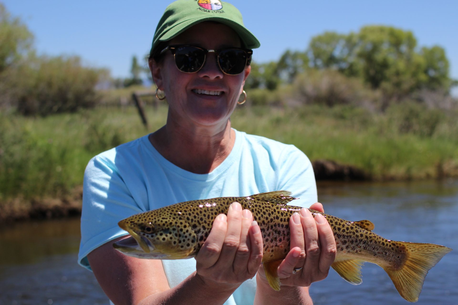 Person in a green cap holding a spotted fish by a riverbank