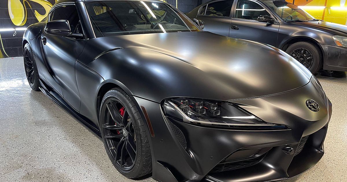 Black matte Toyota Supra parked indoors with another car in the background.