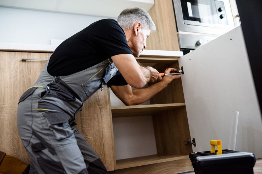 Person in gray overalls installing a cabinet in a kitchen, reaching into an open shelf.