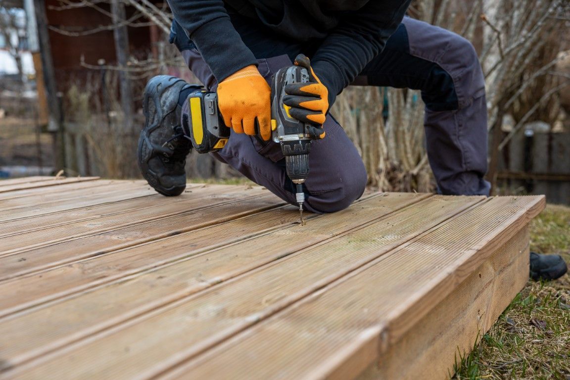 Person using a power drill to fasten wooden deck boards outdoors, wearing orange gloves.