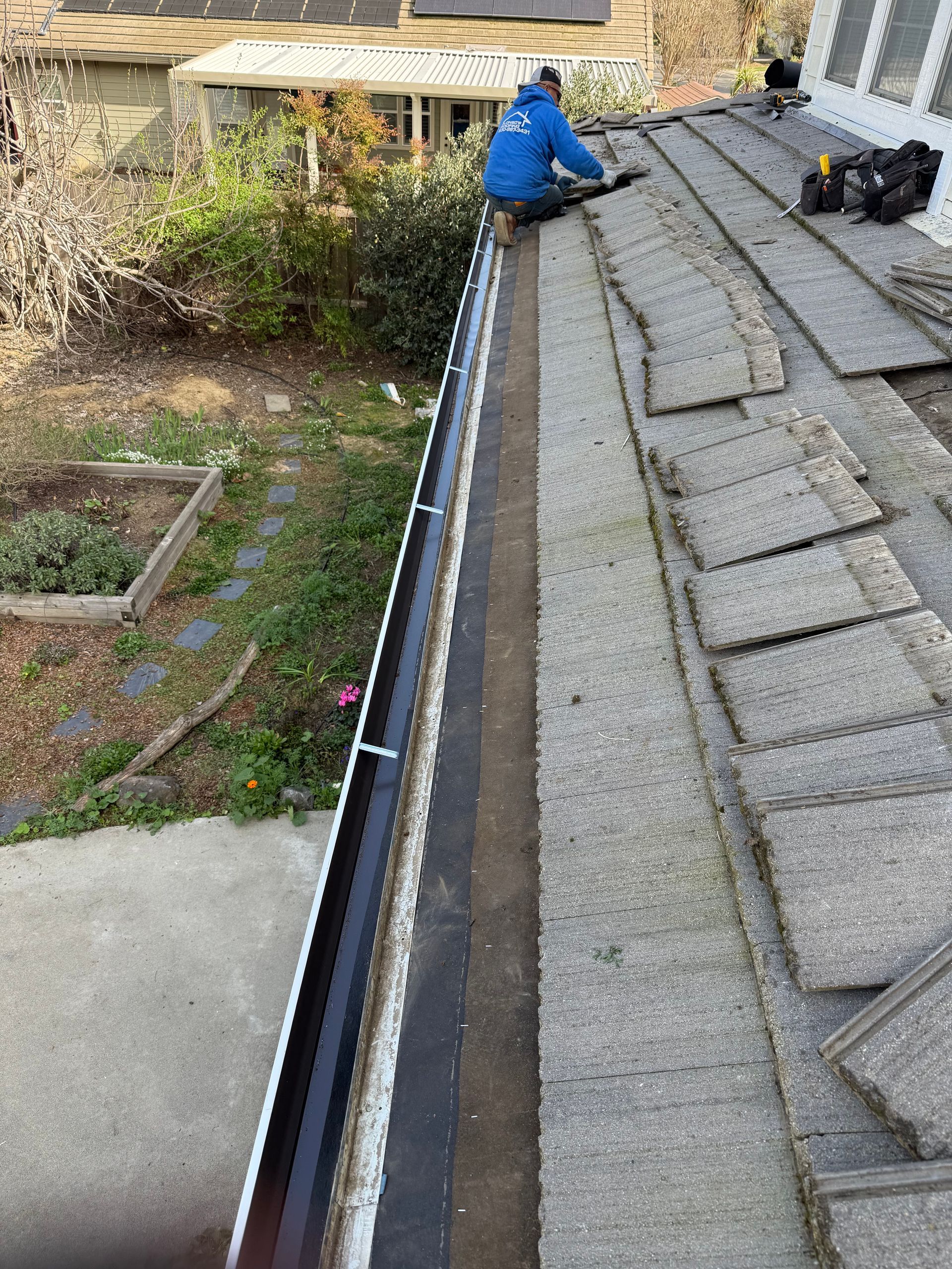 Worker in a blue jacket repairs a roof gutter beside gray shingles and a backyard yard.