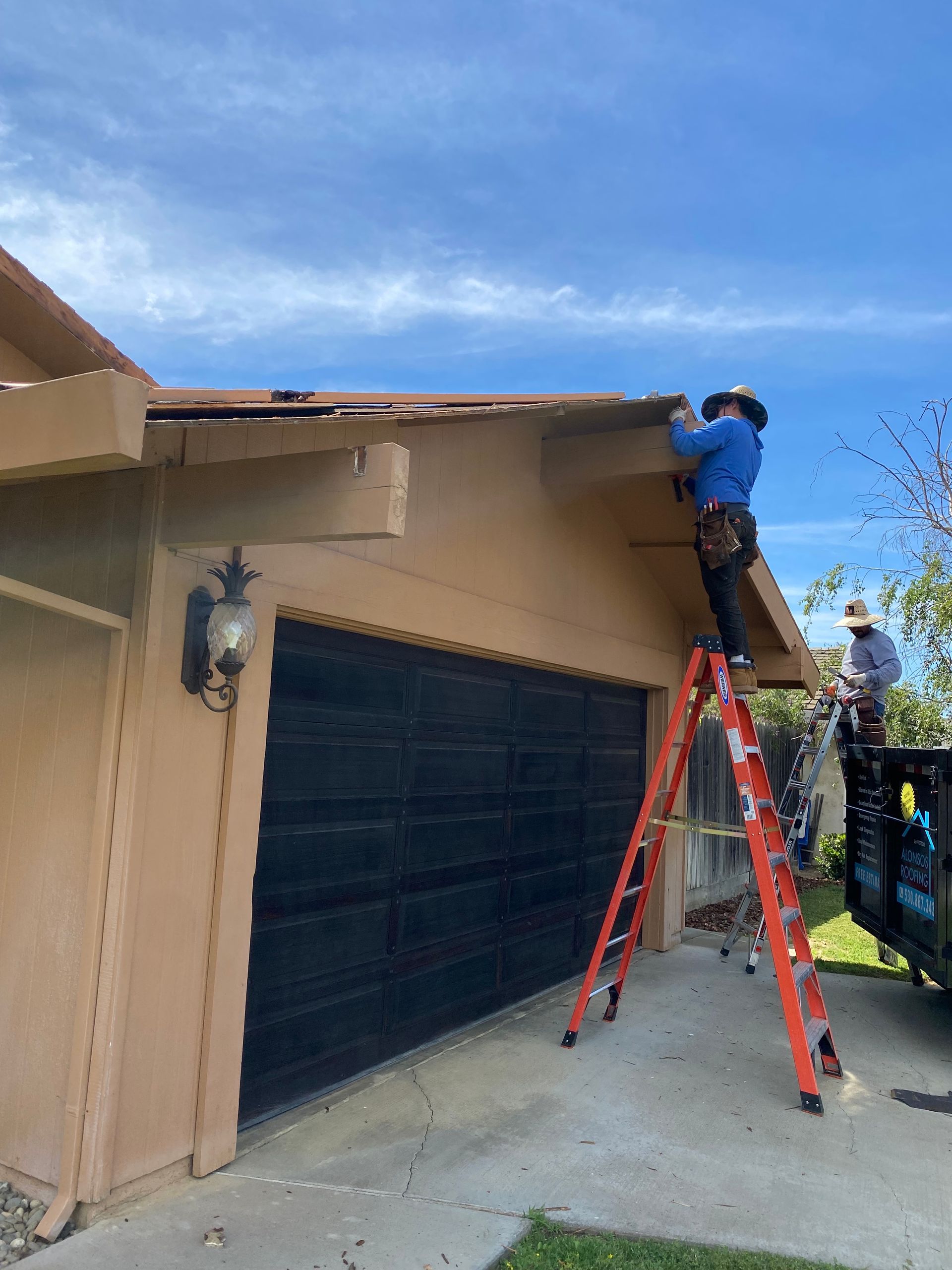 Worker on ladder repairing roof edge above a garage, with another person nearby holding a trash bag.