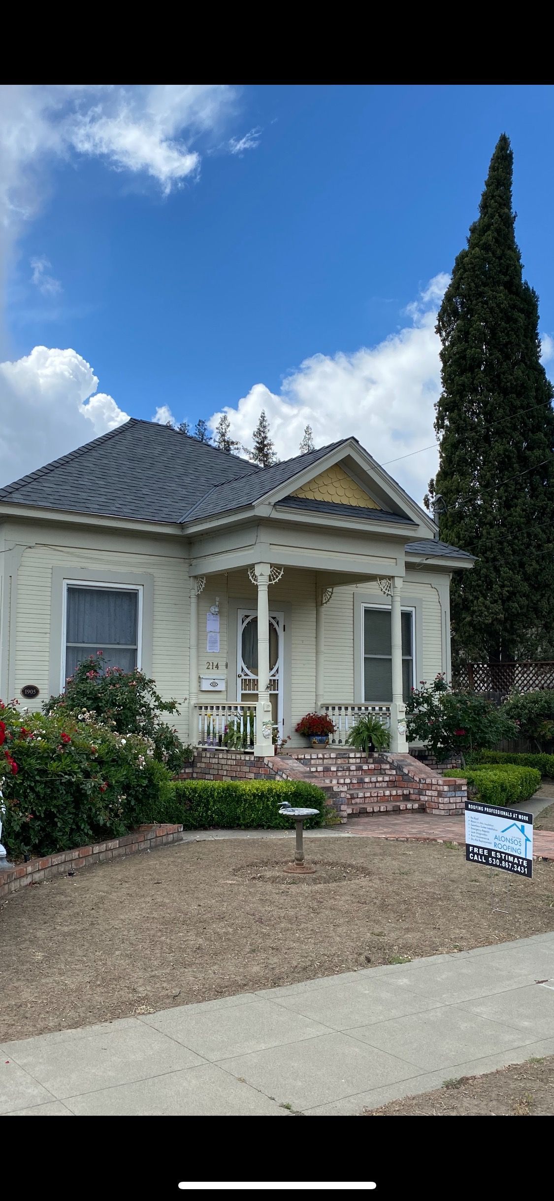Small cream-colored house with a blue roof, front porch, garden, and tall cypress tree under a blue sky.