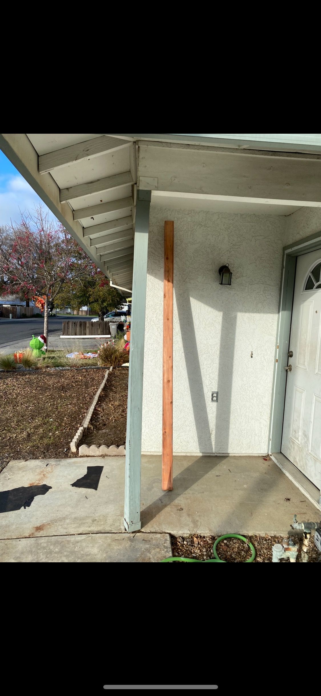 Exterior entryway with white door, wall-mounted light, and wooden post under a covered porch beside a garden.