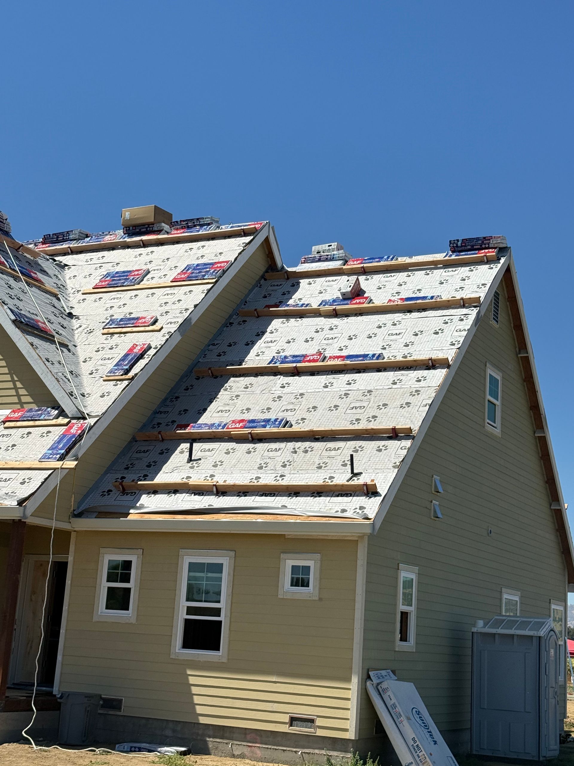 Partially roofed beige house under construction with white underlayment and blue sky