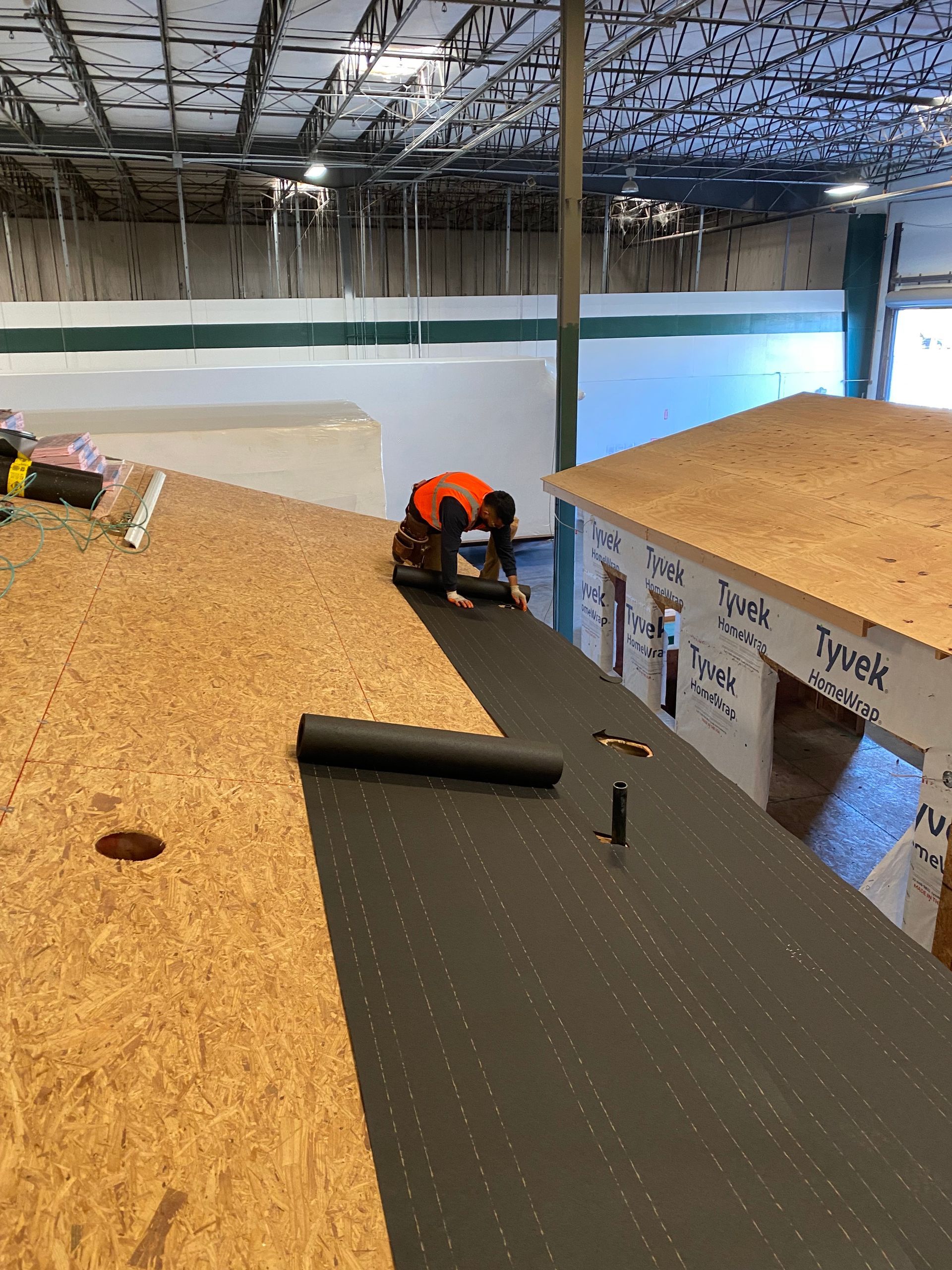 Worker kneeling on a rooftop deck beside a black membrane, with plywood, insulation, and tools nearby.