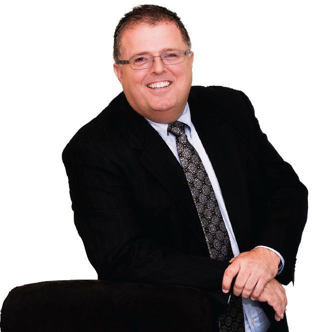 Man in a suit smiles, leaning on a dark chair.