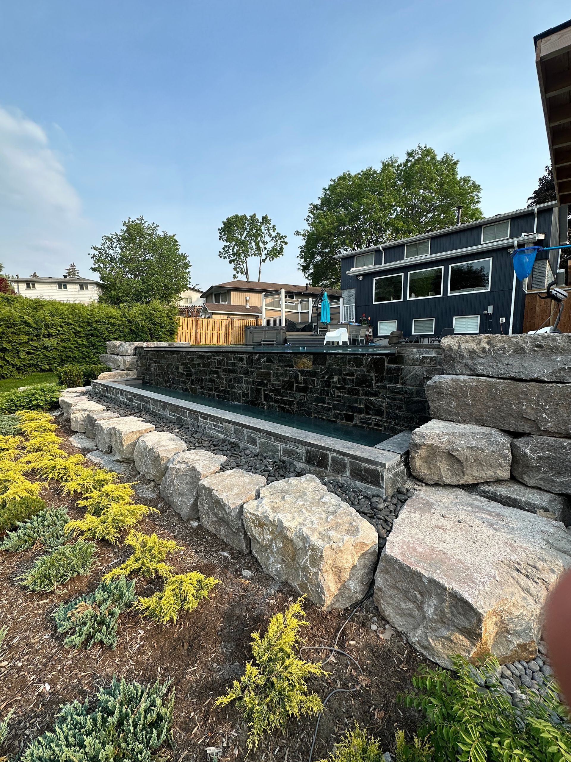 A large rock wall surrounds a swimming pool in a backyard.