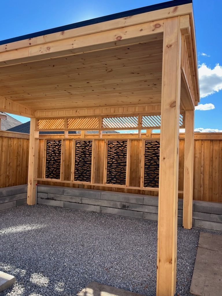 A wooden pergola is sitting in the middle of a gravel yard next to a wooden fence.