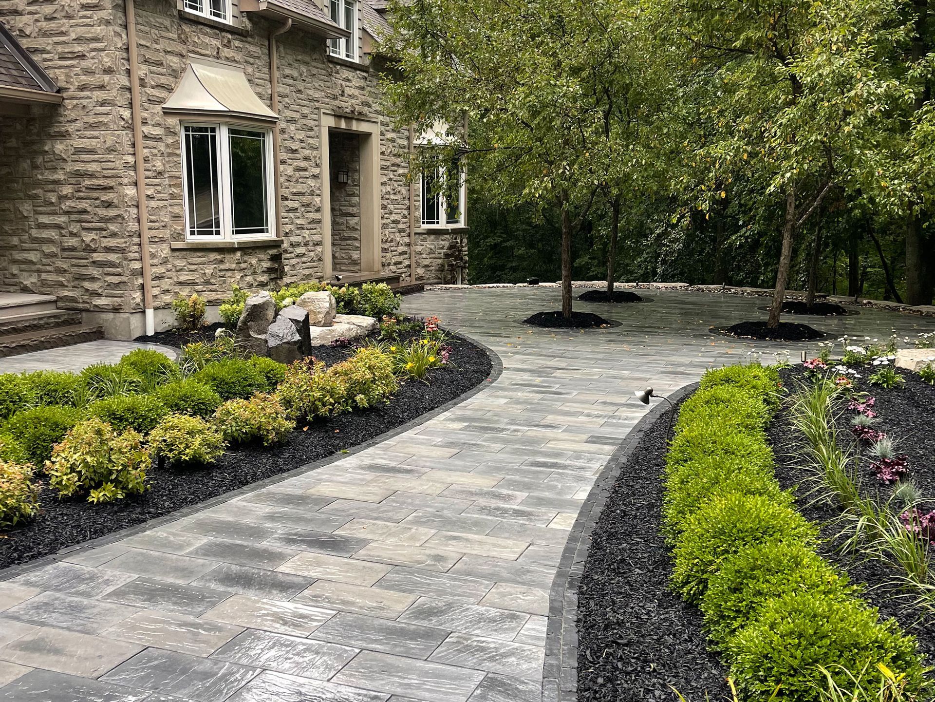 A stone walkway leading to a large brick house surrounded by trees and bushes.