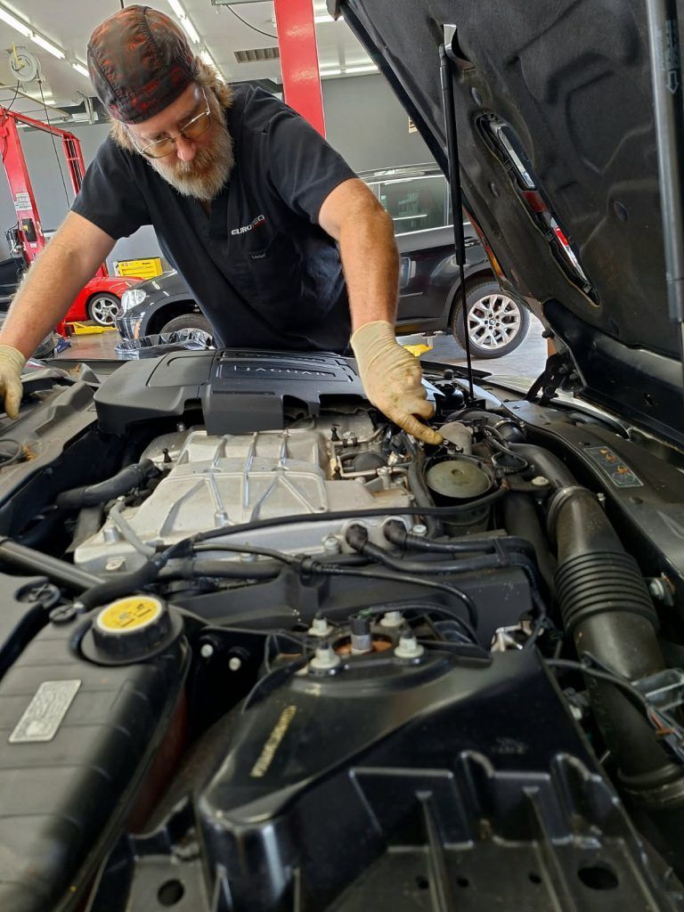 Mechanic working under an open car hood in a garage, inspecting the engine bay.| Eurotech Knoxville
